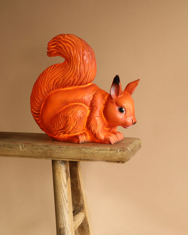 A bright orange Squirrel Lamp with a bushy tail rests on the edge of a wooden bench against a beige background, basking in warm soft light. Its whimsical design evokes the charm of a children's lamp.