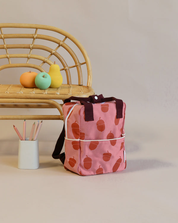 A pink Sticky Lemon Backpack Small with an acorn pattern rests on the floor against a rattan chair, which holds colorful objects and a cup with pencils beside it against a neutral wall.