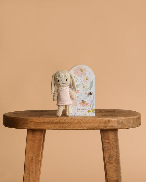 Cuddle + Kind Tiny Hannah The Bunny, hand knit in a pink dress, stands on a wooden table by a floral decorative door against a beige background—a charming, non-toxic doll perfect for thoughtful play.