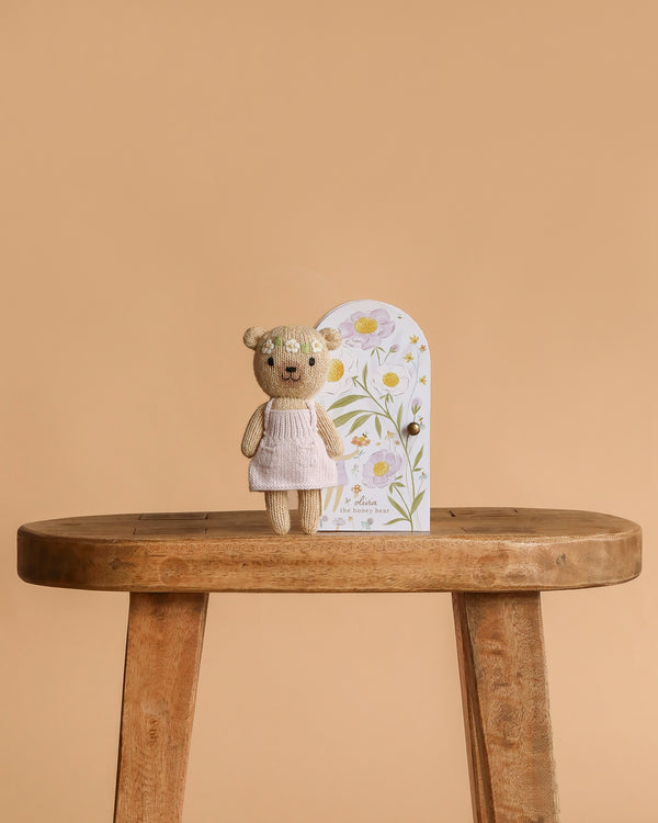 Cuddle + Kind Tiny Olivia the Honey Bear, a hand-knit cotton teddy bear in a pale pink dress, stands on a wooden stool before a floral decorative door, set against a plain beige background.
