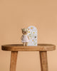 Cuddle + Kind Tiny Olivia the Honey Bear, a hand-knit cotton teddy bear in a pale pink dress, stands on a wooden stool before a floral decorative door, set against a plain beige background.