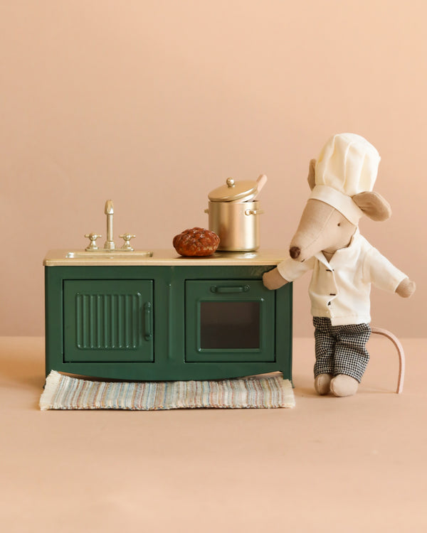 The Maileg Miniature Chef Set features a chef mouse by a green kitchen set, cooking at the stove with a bread loaf and striped rug nearby, set against a beige background.
