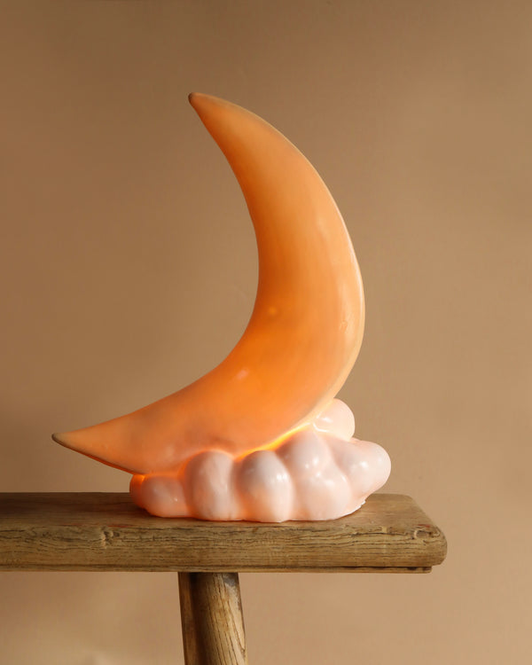 The Crescent Moon Lamp, a crescent-shaped ceramic nursery lamp with a glossy finish, rests on a wooden surface. It emits a warm glow that creates a cozy atmosphere with its whimsical design. The base resembles a cloud-like formation.