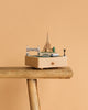 The Paris Music Box, adorned with miniatures of the Eiffel Tower, a bridge, trees, and a boat, is displayed on a rustic wooden stool set against a plain beige background.