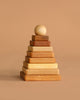 The Natural Square Pyramid Stacker features eco-friendly wooden squares of different sizes stacked in a pyramid with a round wooden ball on top, shown against a plain beige background.
