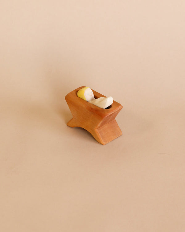 A small handcrafted Ostheimer Baby Jesus sculpture on a matching wooden bed, set against a plain light brown background.