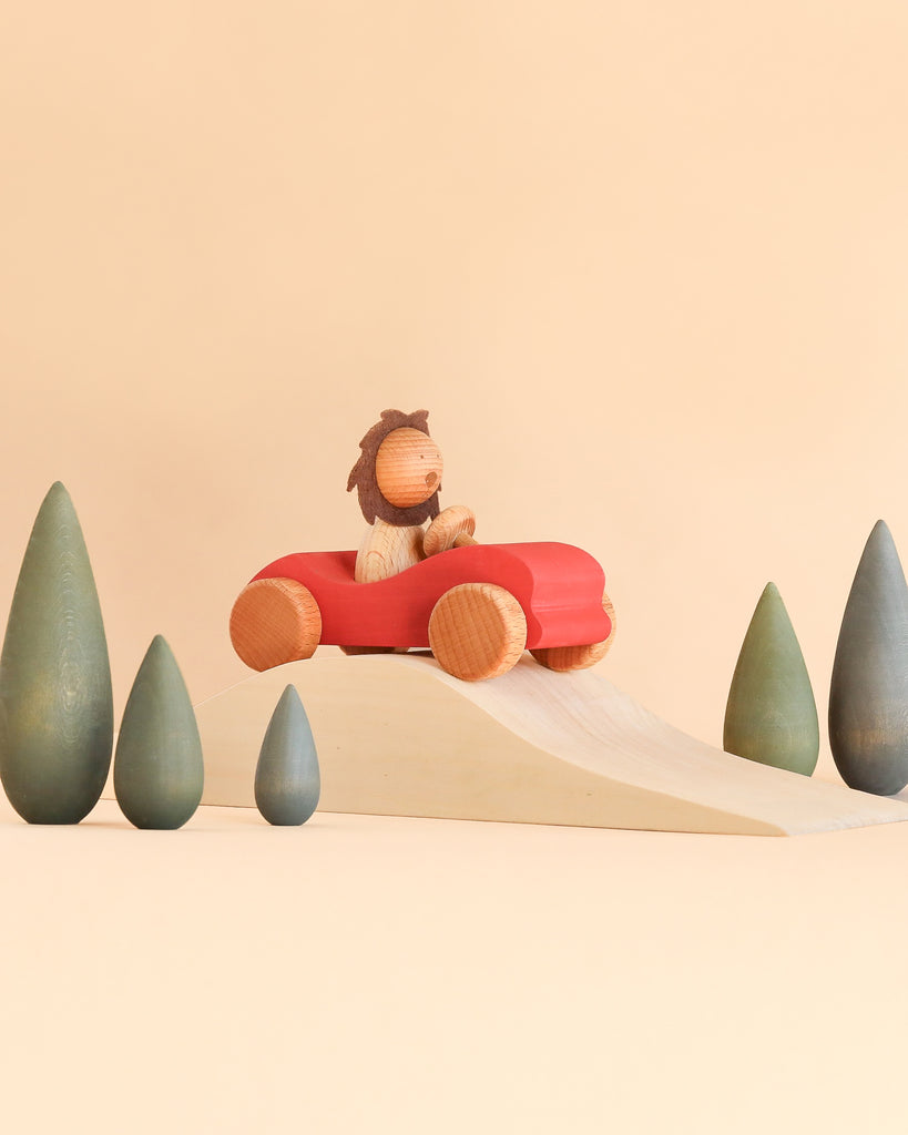 A wooden lion figurine from the Sabo Concept First Journey set drives a red toy car over a wooden ramp, surrounded by small green toy trees designed to develop coordination skills.