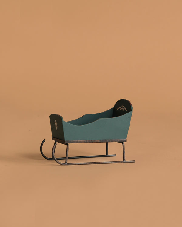The Maileg Sleigh, Mouse - Green is a small, vintage-style green wooden sleigh with black runners and white hand-painted motifs, displayed on a plain light brown background.