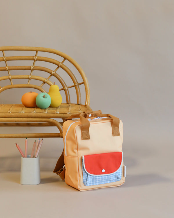 A Sticky Lemon Backpack Small made from recycled PET bottles positioned on the floor next to a wooden bench, upon which rest two apples and a mug containing pencils, against a neutral background.