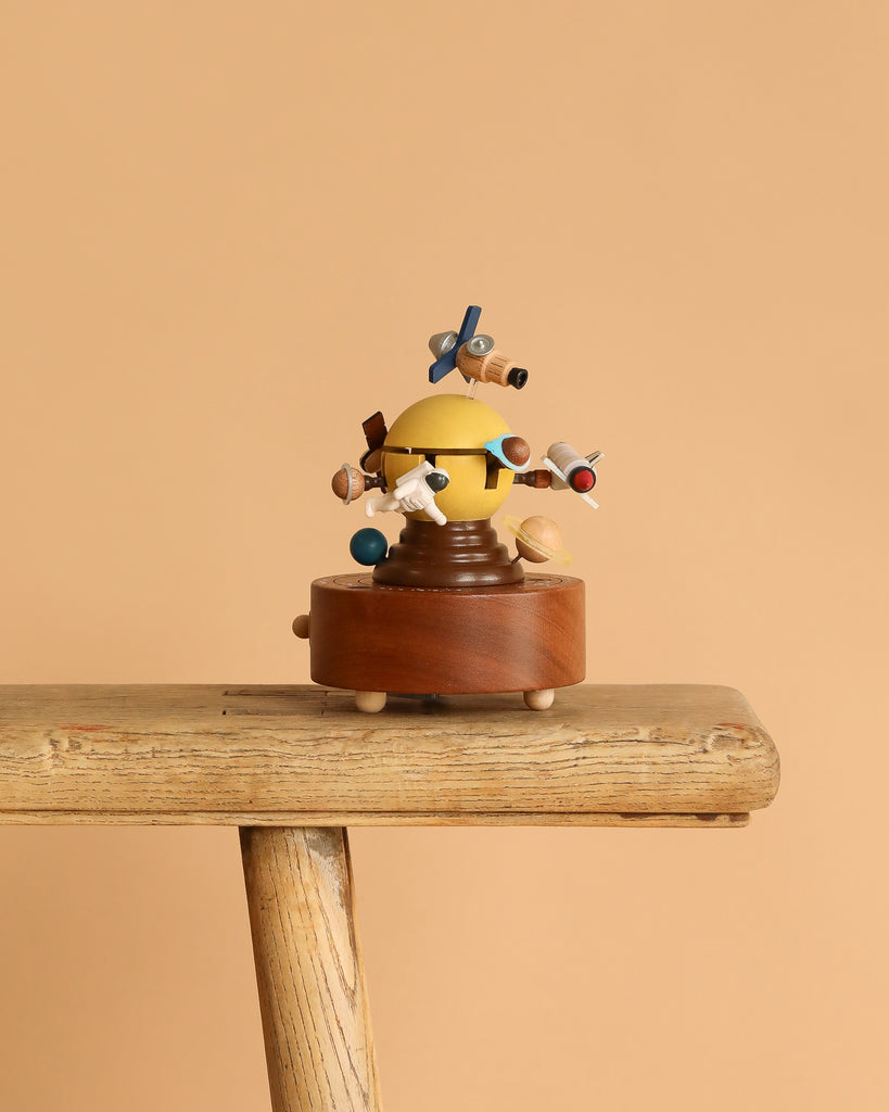 The Space Exploration Music Box by Wooderful Life, with a yellow and blue figure and wooden details, is displayed on a rustic bench against a simple beige background.