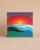 A colorful wooden puzzle featuring a serene landscape design, ideal for developing fine motor skills. The bottom pieces form green and blue waves, the middle has a yellow semi-circle representing the sun, and the top layers depict a gradient sunset with hues of red, orange, purple, and blue. This is the Grimm's Creative Puzzle Landscape Block Set.