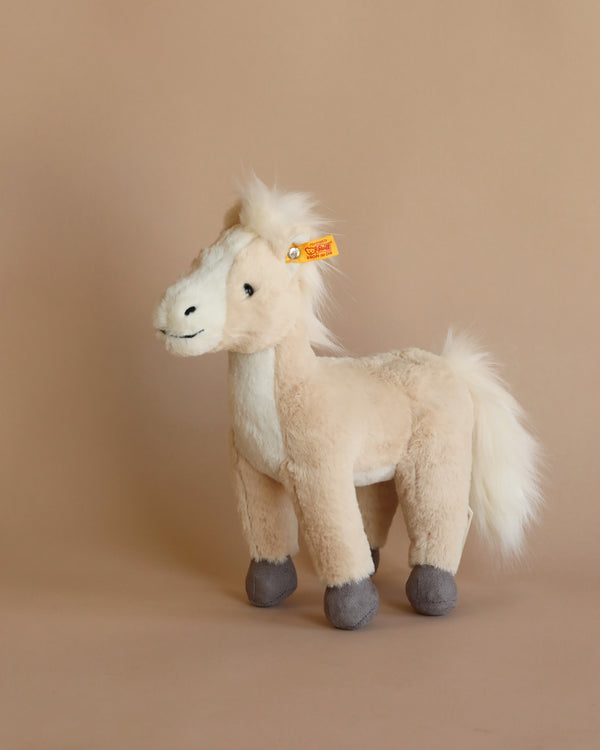 A Stieff brand Gola Standing Horse Plush Stuffed Toy, 11" (as Seen in Barbie) with a cream-colored body, white mane and tail, and grey hooves, featuring the Button in Ear trademark, standing against a light brown background.