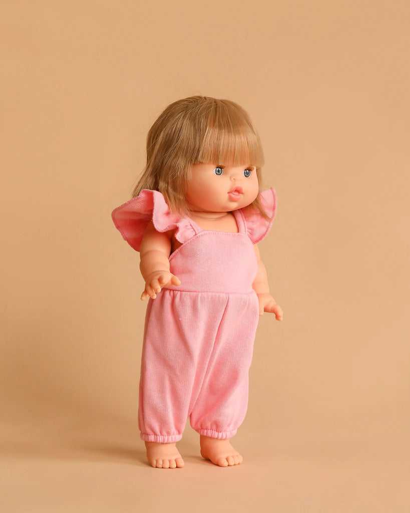 A Minikane doll with straight blonde hair and bangs wears 13” Claudie Overalls in Strawberry Pink cotton, standing barefoot against a beige background.