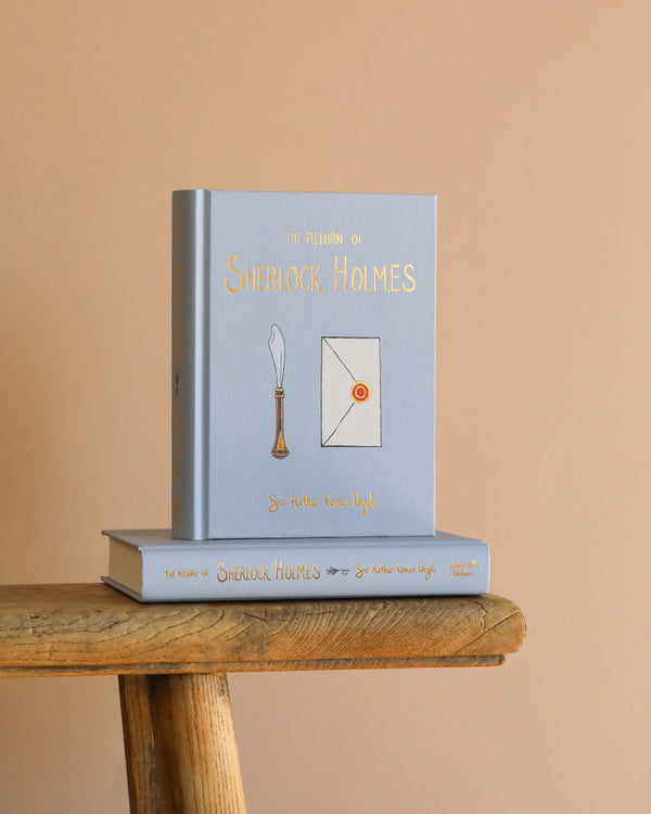 A light blue Collector's Edition hardcover of "The Return of Sherlock Holmes" stands upright on a wooden surface, with another copy flat beneath it, set against a beige background.