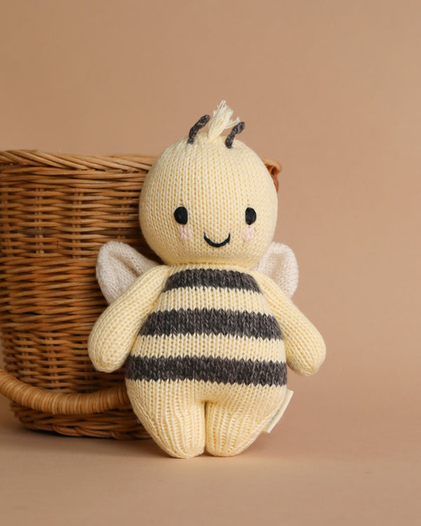 A Cuddle + Kind Baby Bee, crafted from Peruvian cotton yarn, with black and yellow stripes and white wings, sits in front of a small wicker basket on a beige background.