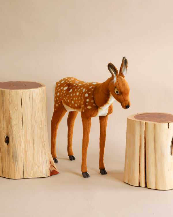 A Large Deer Stuffed Animal - Female with realistic features, characterized by its brown fur and white spots, stands on a beige background. It is positioned between two tree stump props. The stumps are light-colored with a natural wood texture and darker, smooth tops.