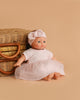 The Minikane Soft Body Doll (11") - Claire with Clothing and Stroller sits in front of a wicker basket on a beige background, wearing an adorable pink dress and matching headband.
