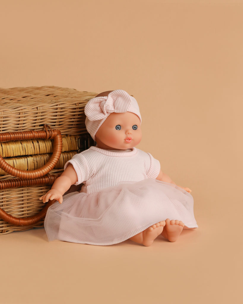 The Minikane Soft Body Doll (11") - Claire with Clothing and Stroller sits in front of a wicker basket on a beige background, wearing an adorable pink dress and matching headband.