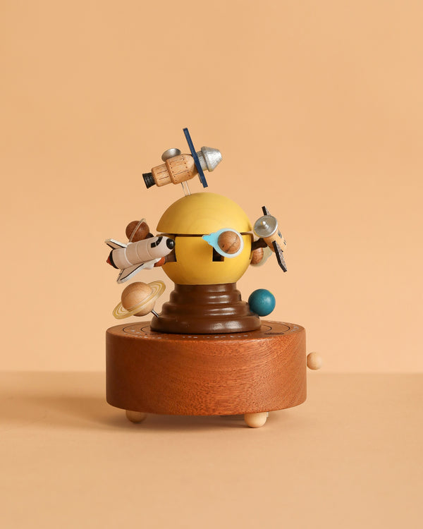 The Space Exploration Music Box by Wooderful Life features planets and spacecrafts orbiting a yellow sphere on a round brown base. This charming wooden decor piece is set against a plain beige background.