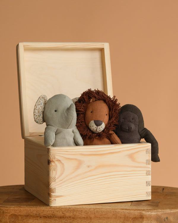 A Maileg Mini Safari Friends Box Set sits open on a wooden surface, displaying three soft stuffed animals—a gray elephant, a brown lion, and a dark brown gorilla—against a plain tan background.
