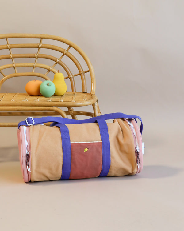 A stylish Sticky Lemon Duffle Bag in soft peach and tan colors sits on the floor in front of a woven chair, on which three colorful pastel toy ducks are placed.