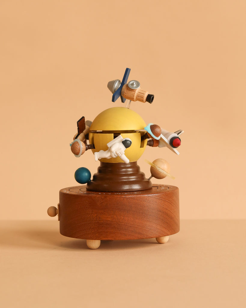 The Space Exploration Music Box features a yellow sphere with planets and space-themed objects orbiting it, all on a round wooden base set against a soft beige background.