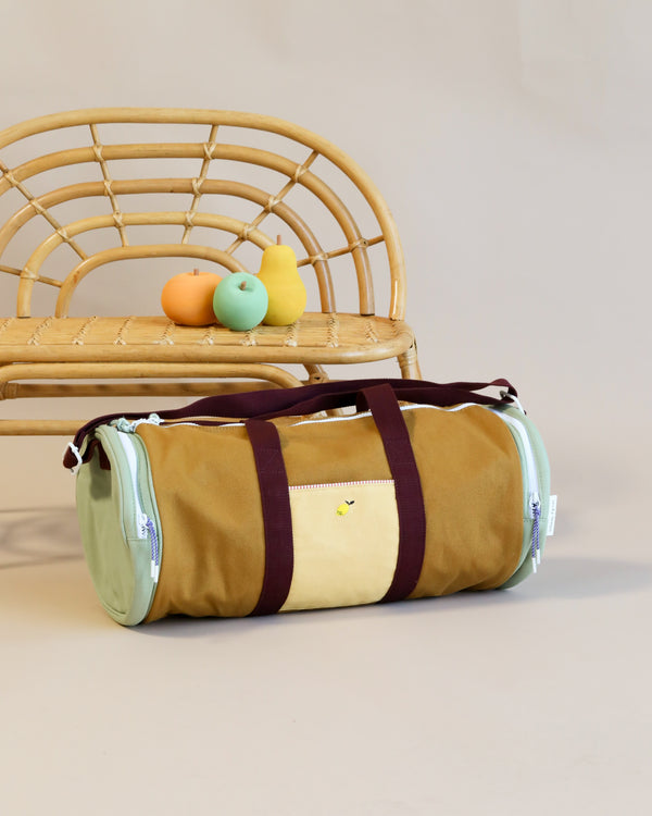 A stylish khaki green Sticky Lemon duffle bag with maroon straps rests in front of a rattan chair, with two colorful toy ducks sitting atop the chair. The background is a neutral cream color.