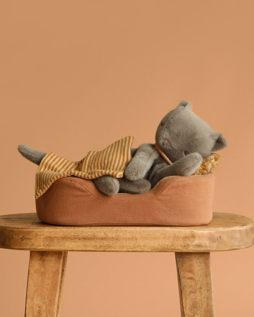 A Maileg Sleeping Kitten in Cozy Bed, plush and gray, rests under a yellow-and-white striped blanket atop a wooden stool with a beige background.