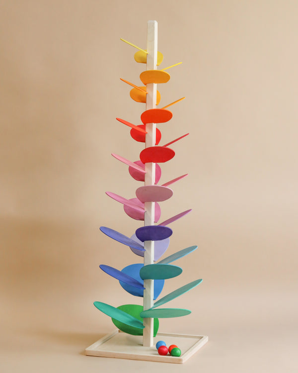 A colorful wooden Giant Magic Wood Marble Tree with flat, elongated paddles in various bright colors, arranged vertically, with wooden marbles at the base ready to descend.