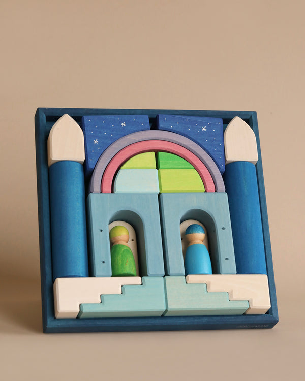 A wooden toy set in pastel colors, part of the enchanting Grimm's Building World Polar Light Block Set, is arranged to resemble a castle. The set includes various geometric shapes and two small wooden figurines standing near the entrance. The background features a night sky with stars, framed in a blue box.