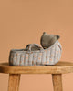 A Maileg Sleeping Kitten in Carry Cot rests in a small, quilted floral basket, set atop a wooden stool against a plain beige background.