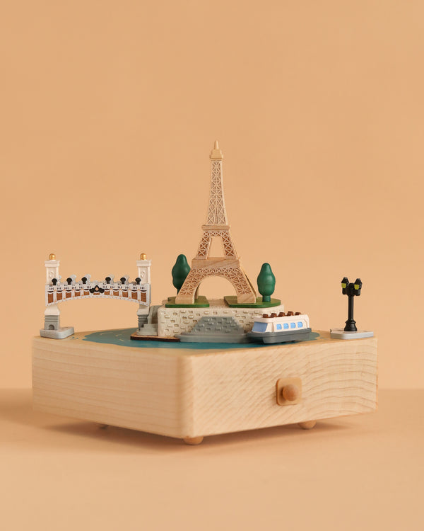 The Paris Music Box displays miniatures of the Eiffel Tower, a riverboat, a bridge, trees, and a street lamp on a plain peach background.