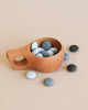 A Wooden Pouring Cup with a handle, filled with smooth, round blue and white stones from the Natural Playbox collection, on a beige background. Additional stones are scattered around the cup.