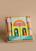 A Grimm's Building World Cloud Play Block Set featuring colorful geometric blocks arranged into a castle-like structure with two cylindrical towers on the sides and arch-shaped blocks in the center. Made from solid wood, it includes two peg dolls placed inside the arch openings, one turquoise and one orange, for imaginative free building.