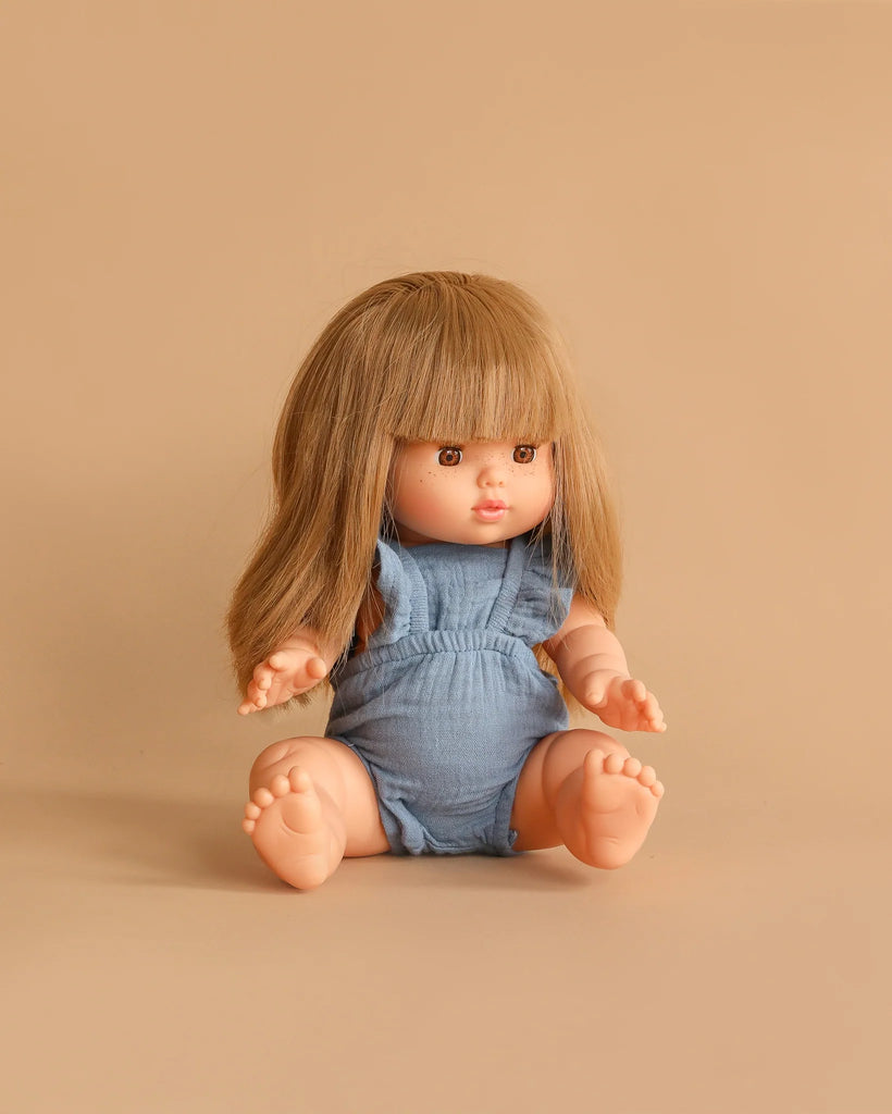 A Minikane doll wears the Lou Retro Romper in Cotton Gauze - Blue Denim, sitting on a beige surface with a matching beige background. The doll has straight, long blonde hair and bangs.