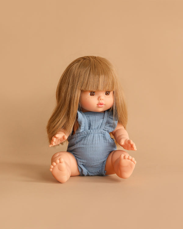 The Minikane Baby Doll (13") - Nina with Clothing features long brown hair and bangs, dressed in a blue romper, and is anatomically correct. Made in Spain, she sits on a beige surface against a neutral background.