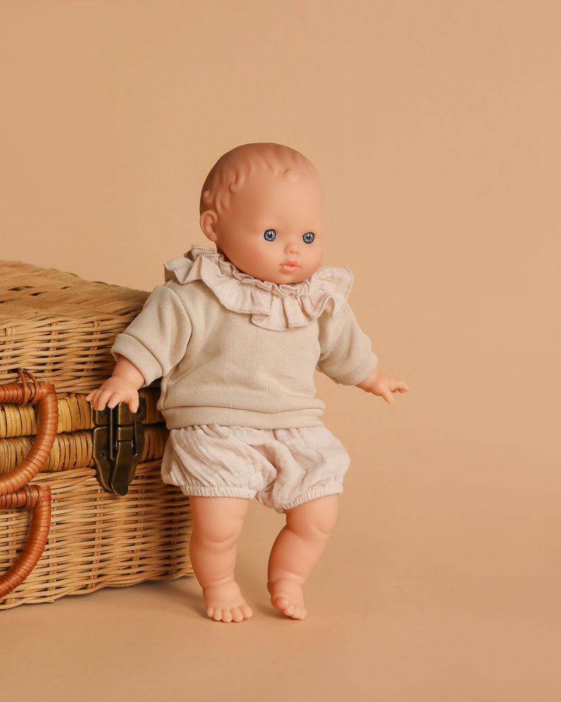The Minikane Soft Body Doll (11") - Claire with Clothing features blue eyes and a light beige ruffled outfit, posed by a wicker basket with a leather latch—a perfect bedtime companion in a cozy beige setting.