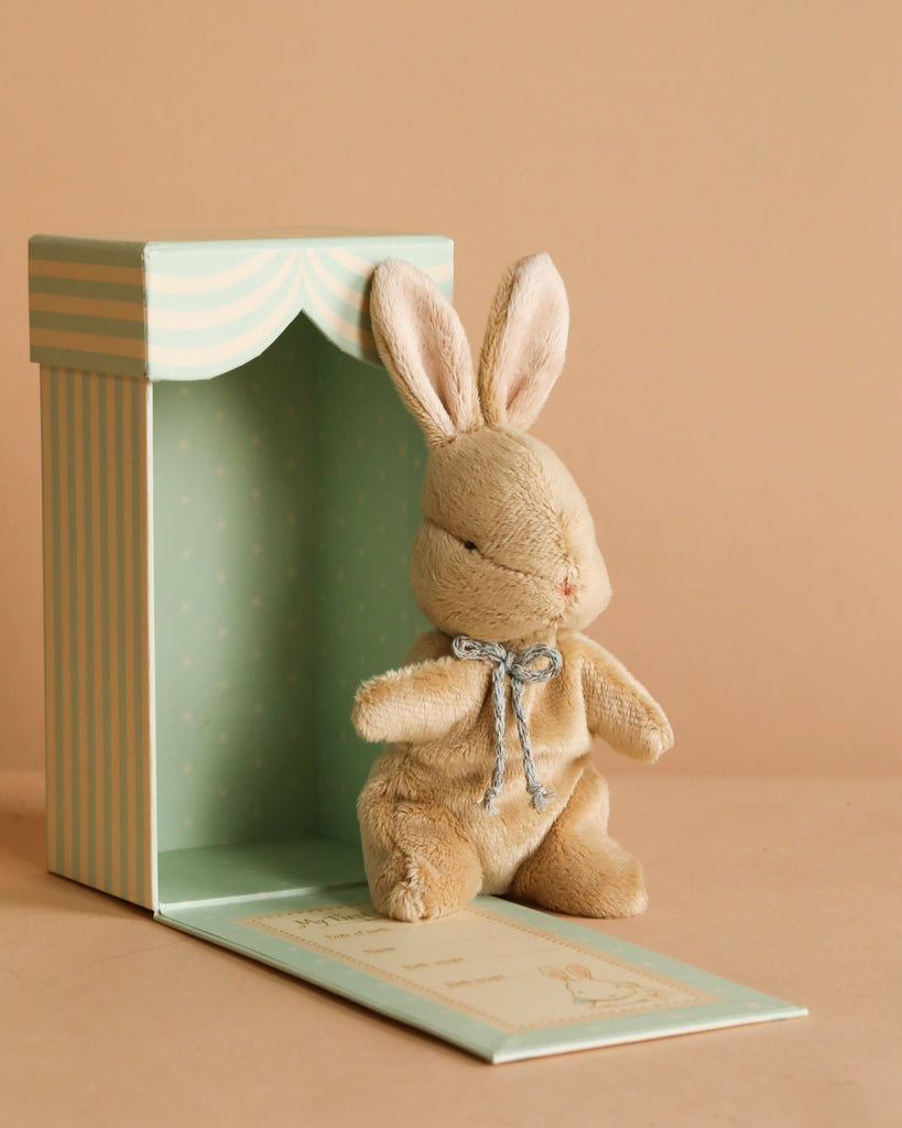 The Maileg My First Bunny - Brown, a small plush bunny with a ribbon bow, sits in front of an open mint green striped gift box on a beige background—an adorable surprise for any occasion.