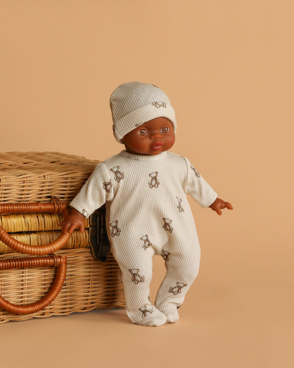 The Minikane Soft Body Doll (11") - Ondine with Clothing, featuring a dark skin tone, wears a cream hat and teddy bear print onesie. It stands next to a wicker basket against a light brown background—an ideal bedtime companion.