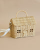 The Olli Ella Rattan Casa Clutch is a wicker purse shaped like a toy house, featuring roof, windows, and door details. Made from natural rattan, it includes a top handle and an attached white shoulder strap. The background has a solid beige color.