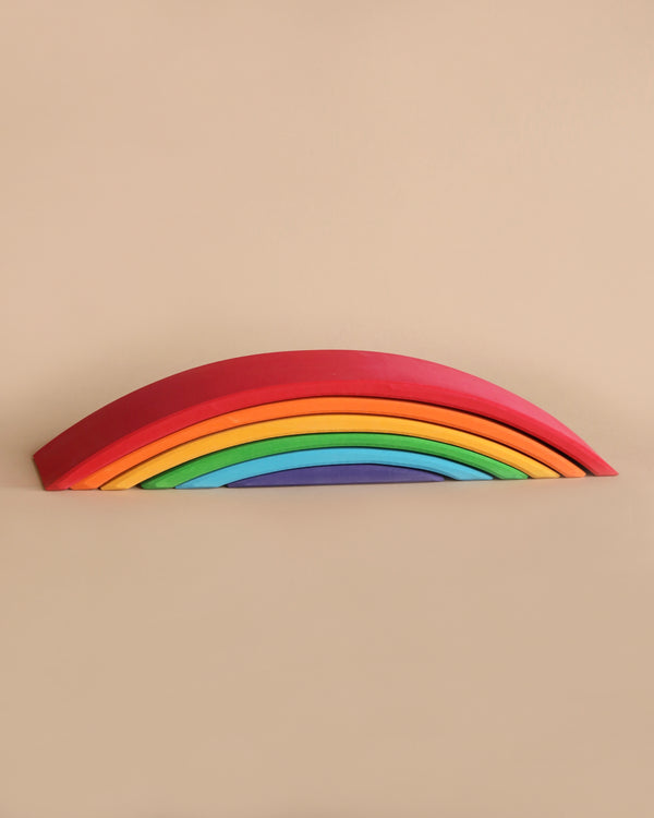 A minimalist image features a non-toxic Grimm's Rainbow Bridge against a beige background. The toy consists of arch-shaped pieces stacked on top of each other, arranged in the order of a natural rainbow: red, orange, yellow, green, blue, and purple.