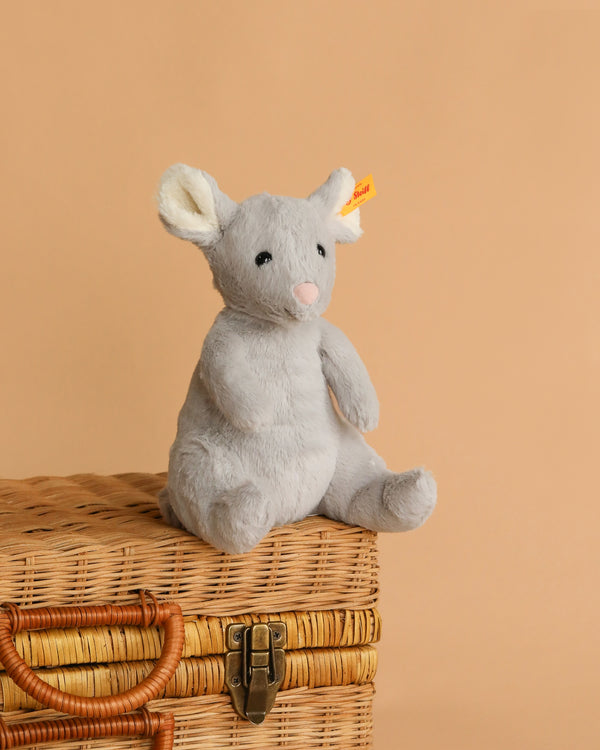 The Steiff Cheesy Mouse Stuffed Animal, featuring white ears, a pink nose, and bean bag stuffing, sits on a closed woven wicker suitcase against a plain beige background.