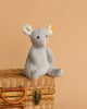 The Steiff Cheesy Mouse Stuffed Animal, featuring white ears, a pink nose, and bean bag stuffing, sits on a closed woven wicker suitcase against a plain beige background.