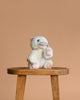 The Olli Ella Dinkum Cat Lux - White, a posable plush kitten with pink ears and paws, sits on a wooden stool against a beige background, ready for play.