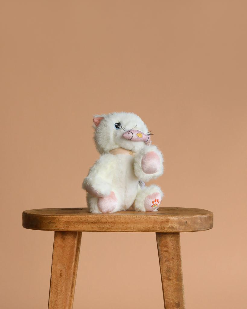 The Olli Ella Dinkum Cat Lux - White, a posable plush kitten with pink ears and paws, sits on a wooden stool against a beige background, ready for play.