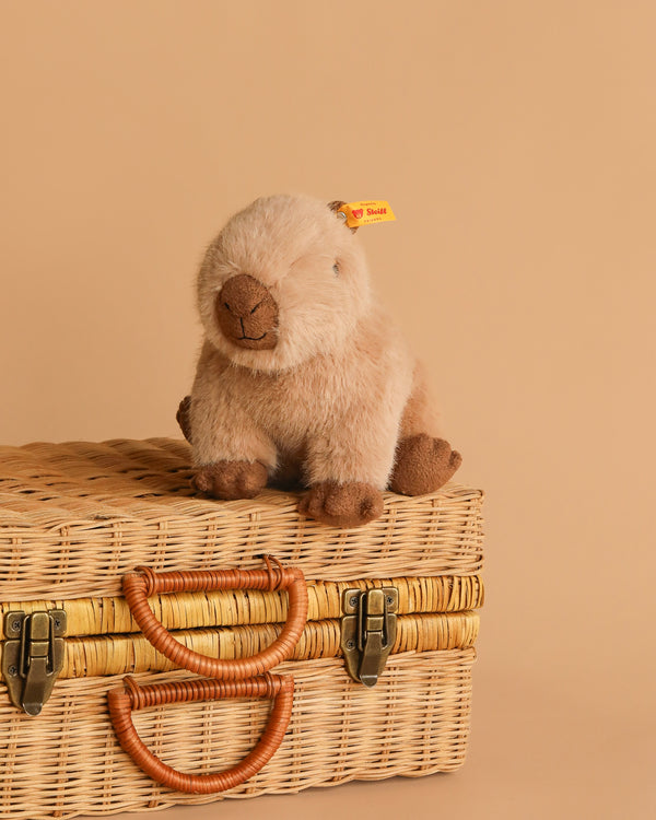 A Steiff Chip Capybara plush toy with a yellow Steiff tag sits atop a closed wicker picnic basket with leather handles and metal clasps, set against a beige background.