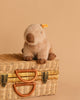 A Steiff Chip Capybara plush toy with a yellow Steiff tag sits atop a closed wicker picnic basket with leather handles and metal clasps, set against a beige background.