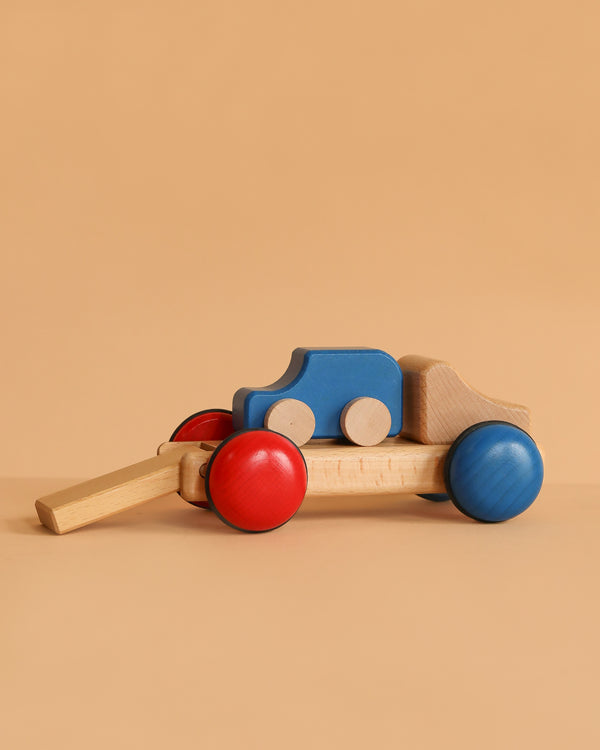 The Fagus Wooden Car Transporter - Mini Series, featuring red and blue wheels and a blue body, is displayed on a beige surface with a beige background.