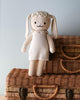 Cuddle + Kind Hannah the Bunny, hand-knit from premium cotton yarn and dressed in a cream dress with a flower crown, sits on two stacked wicker suitcases against a light blue wall.