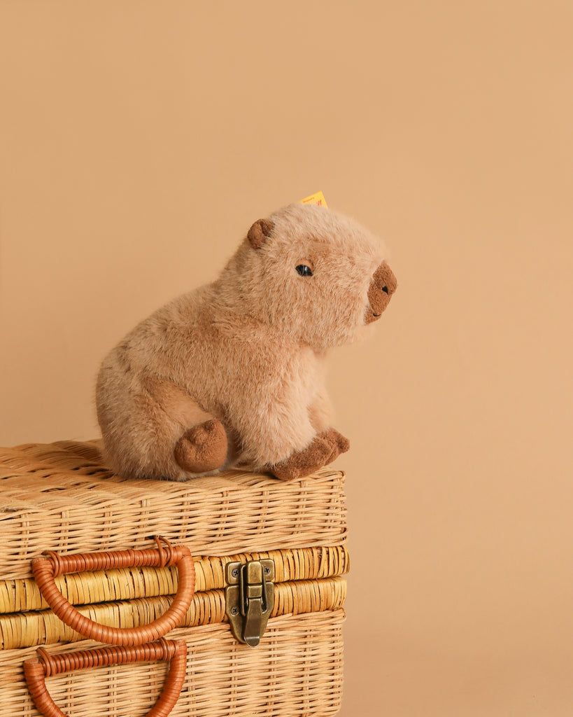 The Steiff Chip Capybara plush toy sits adorably on a closed wicker picnic basket with a plain beige background.